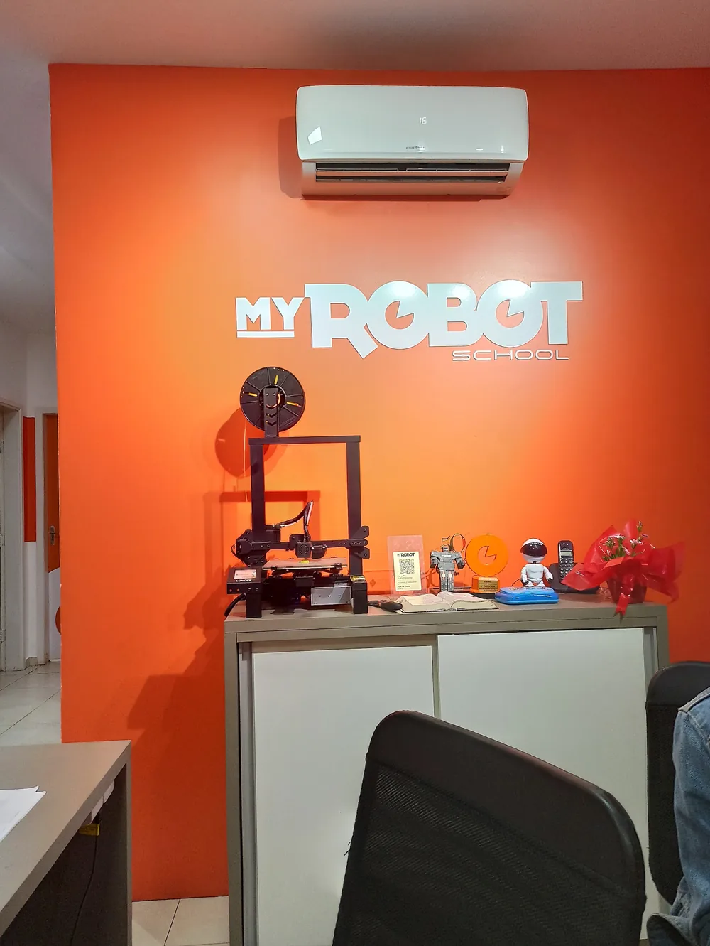 Parede laranja da escola com o logotipo My Robot School e a impressora 3D sobre o balcão