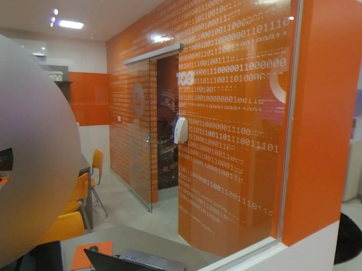 Sala de aula da My Robot Campo Grande vista pelo vidro: parede laranja com código binário, cadeiras e notebooks nas bancadas