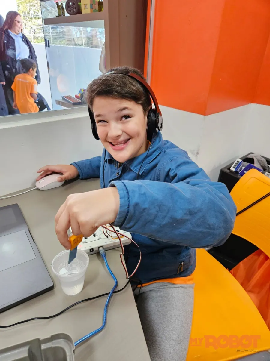 Criança de fone sorrindo enquanto faz um experimento com copo d'água e sensor na aula de Onebot